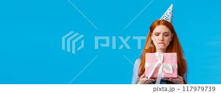 Tempting moment of unwrapping gift. Cute glamour redhead teenage girl biting lip eager open cute birthday present, wearing silly cap, celebrating b-day standing blue background curious Tempting moment of unwrapping gift. Cute glamour redhead teenage girl biting lip eager open cute birthday present, wearing silly cap, celebrating b-day standing blue background curious 117979739
