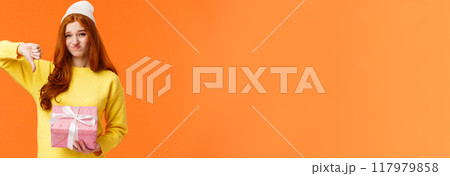 Skeptical and awkward cute redhead picky girl dont like christmas gift from aunt, show thumb-down, grimacing with uncomfortable judgemental expression, holding pink ugly present, orange background 117979858