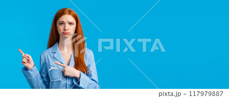 Regret, bad mood and advertising concept. Sad unhappy redhead girl in nightwear, sulking and frowning express disappointment or jealousy, pointing fingers right, standing blue background 117979887