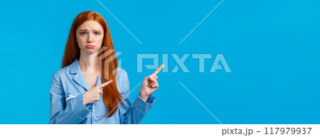 Sad sulking cute teenage girl with red hair asking parents something on discount, pouting frowning making offended jealous expression, standing blue background in nightwear, pointing left 117979937