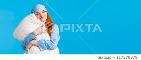 Beauty, tenderness concept. Happy feminine cute redhead teenage girl in nightwear, sleep mask, close eyes and smiling delighted, finally went bed after long day, hugging pillow, blue background 117979979