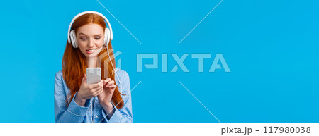 Waist-up portrait indecisive, unsure picky cute redhead female in nightwear, biting lip and looking tempting with desire smartphone screen, send risky text, listen music headphones 117980038