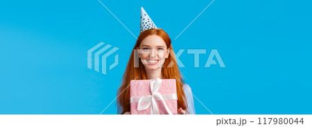 Waist-up portrait cheerful happy, pretty redhead, ginger girl celebrating birthday, receive cute pink wrapped present, wearing b-day cap and smiling, having fun at party, blue background 117980044