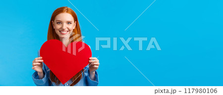 Happy, cheerful and lovely, tender caucasian woman holding big red heart valentines card and smiling, giving her love and care, congratulate boyfriend with their romantic date, blue background 117980106