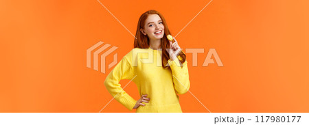 Waist-up portrait lovely redhead girl holding tasty yellow macaron near mouth and smiling joyfully, like eating sweets, buy dessert for romantic valentines date, posing cheerful orange background 117980177