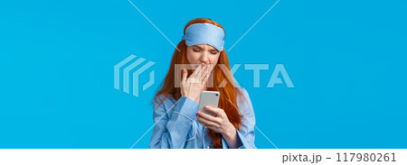 Gadgets addication, lifestyle and beauty concept. Lovely redhead girl going sleep and scrolling feed smartphone, yawning squinting feeling tired, wearing pyjama and sleep mask, blue background Gadgets addication, lifestyle and beauty concept. Lovely redhead girl going sleep and scrolling feed smartphone, yawning squinting feeling tired, wearing pyjama and sleep mask, blue background 117980261