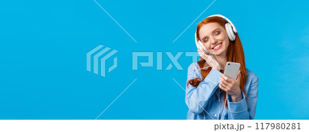 Tenderness, women and lifestyle concept. Cheerful tender redhead female in pyjama, tilting head enjoying listening music, holding smartphone and smiling as touching earphones from delight Tenderness, women and lifestyle concept. Cheerful tender redhead female in pyjama, tilting head enjoying listening music, holding smartphone and smiling as touching earphones from delight 117980281