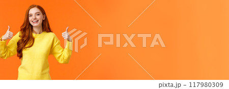 Holidays, gestures, people concept. Cheerful cute redhead girl smiling and showing thumbs-up recommend product, give positive reply, agree or like something, nod agreement, orange background 117980309