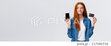 Shopaholic cant hold herself want buy something online. Eager and excited redhead girl see tempting price internet store, biting lip thrilled stare smartphone display, hold credit card 117980338