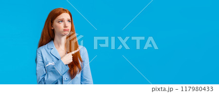 Envy and sad sulking moody redhead teenage girl in nightwear, looking and pointing upper left corner unsatisfied, jealous expression, seeing something she wants, feeling uneasy, blue background 117980433