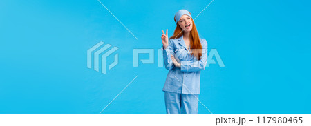Saying cheeze. Cheerful and cute redhead upbeat girl in glamour pyjama, fancy nightwear having sleepover showing peace sign and smiling excited, having upbeat lucky morning, standing blue background 117980465