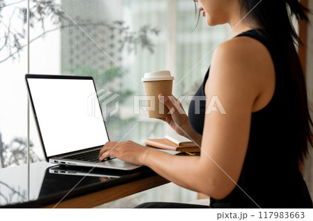 A woman working remotely from a minimalist coffee shop, using a laptop while having coffee. A woman working remotely from a minimalist coffee shop, using a laptop while having coffee. 117983663