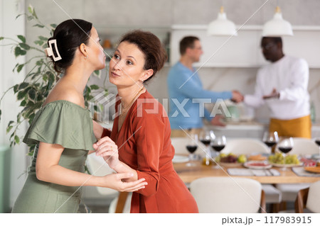 Women greet each other with kiss on cheek before festive dinner. Men shaking hands in background 117983915