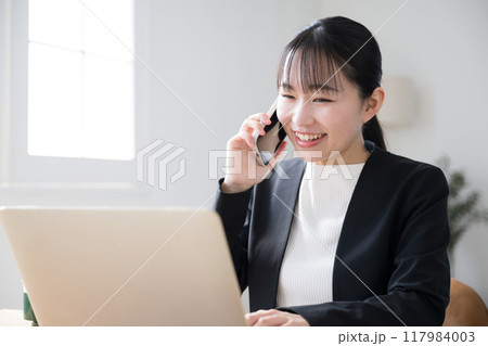 パソコンを操作しながら電話をするビジネスシーンの女性　笑顔で画面に向かって話す	 117984003