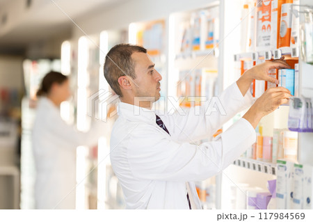 Young male pharmacist putting medical product on shelf in drugstore 117984569