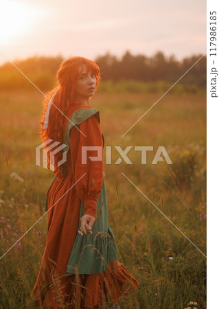 A shepherdess woman with red hair and a green dress sits in a field of flowers. 117986185