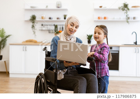Mother in wheelchair with daughter using laptop in modern kitchen 117986875