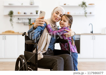 Muslim mother in wheelchair taking selfie with daughter at home 117986877