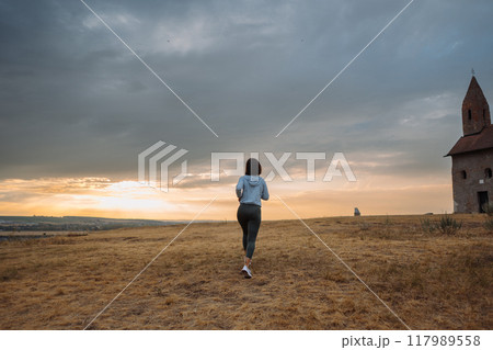Woman Jogging by Historic Church at Sunset in Scenic Countryside 117989558