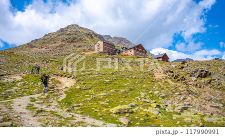 Hikers trek along a path towards Breslauer Hut, nestled in the stunning Otztal Alps of Austria, surrounded by rocky terrain and lush greenery under a bright blue sky with scattered clouds. 117990991