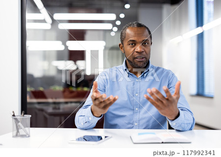 Businessman sitting at desk in modern office, engaged in serious video call. Communicating confidently, looking at camera with open gestures. Businessman sitting at desk in modern office, engaged in serious video call. Communicating confidently, looking at camera with open gestures. 117992341