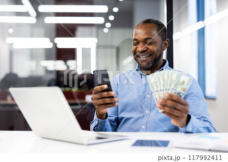 Smiling businessman sitting in modern office, holding cash and phone, symbolizing success and financial prosperity. Laptop on desk suggests productive work environment and technological engagement. 117992411