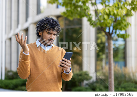 Young man feeling frustrated while looking at phone outside modern office building. He gestures with confusion, expressing annoyance. Wearing brown sweater, standing in urban environment under tree. Young man feeling frustrated while looking at phone outside modern office building. He gestures with confusion, expressing annoyance. Wearing brown sweater, standing in urban environment under tree. 117992569