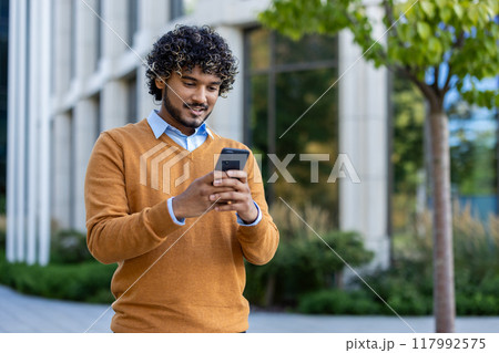 Young man enjoying outdoor work break, standing with phone in hand. Wearing casual sweater, he engages with technology in urban environment surrounded by nature, reflecting modern lifestyle 117992575