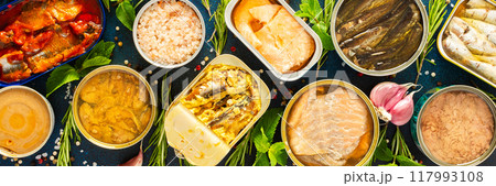 Enjoy an assortment of canned fish including salmon, mackerel, sprat, sardine, tuna, herring, and fish pate, displayed in open tins with spices and herbs on a blue background, banner 117993108