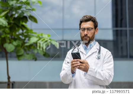 Male doctor standing outdoors wearing white coat and stethoscope, using phone. Medical professional looking serious, focused on digital device in urban setting. Concept of healthcare, technology 117993571