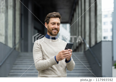 Confident man holding phone, smiling as he stands in front of corporate building. Represents business connectivity, technology, and communication. Ideal image for depicting professional lifestyle Confident man holding phone, smiling as he stands in front of corporate building. Represents business connectivity, technology, and communication. Ideal image for depicting professional lifestyle 117993592