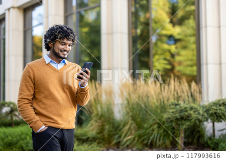 Confident man standing outside modern office building, holding phone. Wearing casual orange sweater, enjoying sunny day. Reflection of trees seen on glass windows, creating calm serene atmosphere. 117993616