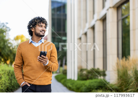 Hispanic businessman in orange sweater holding phone while walking outside office building. Smiling confidently and looking forward, showcasing modern urban work environment , professional lifestyle. Hispanic businessman in orange sweater holding phone while walking outside office building. Smiling confidently and looking forward, showcasing modern urban work environment , professional lifestyle. 117993617