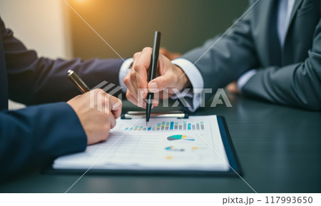 Professionals in business setting analyzing data charts, pen in hand pointing at paper, representing collaboration, decision-making, data analysis in corporate environment 117993650