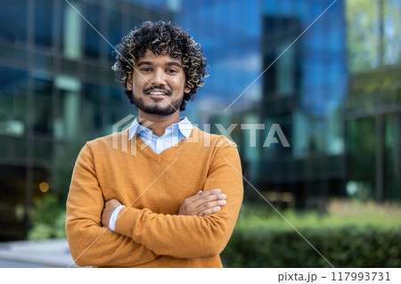 Confident young businessman standing outside modern office building. Business professional smiling with arms crossed, embodying success and positivity. Urban background 117993731