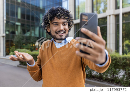 Cheerful man using phone to take selfie outside contemporary office building. Wearing orange sweater and blue shirt, he exudes happiness and confidence. 117993772