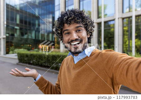 Cheerful businessman taking selfie outdoors, expressing happiness and confidence. Standing near modern office building with inviting smile and relaxed demeanor. 117993783