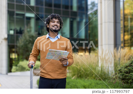 Young businessman holding map and suitcase outside modern office building. He looks ready to embark on travel journey, exuding confidence and curiosity. Perfect portrayal of urban exploration. 117993797