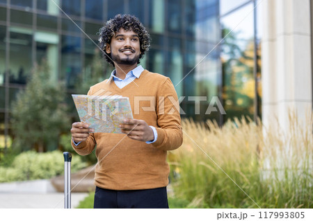 Confident man stands outdoors holding map, looking away with a smile. Dressed in casual attire, he exudes positivity. Urban setting in background suggests travel, exploration. Perfect for travel 117993805