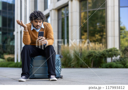 Young man looking at phone with confusion sitting on suitcase outside modern office building. Business professional appearing frustrated while waiting, balancing technology usage in urban setting. 117993812