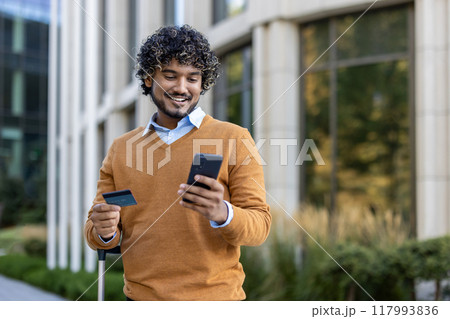 Smiling man stands outside modern building holding phone and credit card. Wearing casual sweater, he appears engaged in online shopping or digital banking, combining technology and convenience 117993836