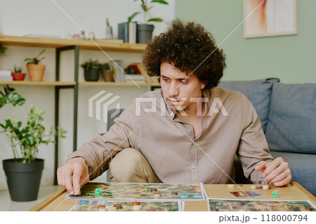 Medium shot of young gen Y biracial man sitting at coffee table in living room at home and playing modern board game 118000794
