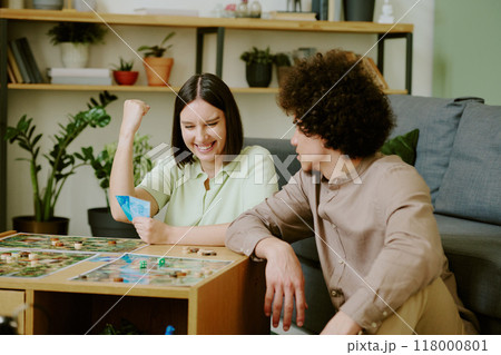 Cheerful young Caucasian woman sitting at coffee table playing board game with her boyfriend and winning round 118000801