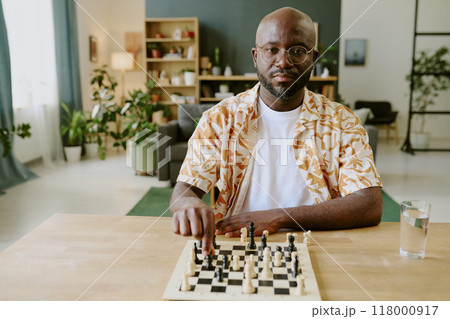 Medium portrait of young African American man wearing casual clothes sitting at desk in living room and looking at camera while solving chess problem 118000917