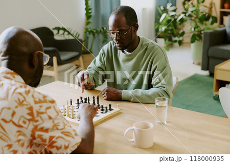 High angle over shoulder shot of two young African American men sitting at table in front of each other in living room playing chess High angle over shoulder shot of two young African American men sitting at table in front of each other in living room playing chess 118000935