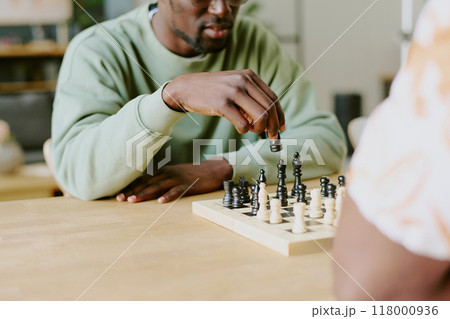 Young Black man sitting at wooden table playing chess game with his unrecognizable friend Young Black man sitting at wooden table playing chess game with his unrecognizable friend 118000936