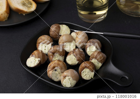 Escargot bourguignon with butter and garlic. Traditional French dish made from snails, served with baguette and white wine. Selective focus, close-up, black background. 118001016