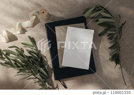 Mediterranean summer wedding stationery mock-up. Blank greeting card invitation, craft envelope and black diary. Olive branches, silk ribbon on beige limestone background in sunlight. Moody flat lay 118001350