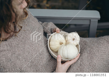 Young girl, woman wearing brown wool sweater holding bowl of little white pumpkins for autumn decorations. Fall festive still life. Halloween, Thanksgiving holiday. Cozy lifestyle. Blurred background 118001351