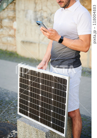 Cropped photo of man charging phone by portable solar panel. Young male using solar battery charger for mobile devices. Concept of alternative energy resources and sustainable lifestyle. 118004883